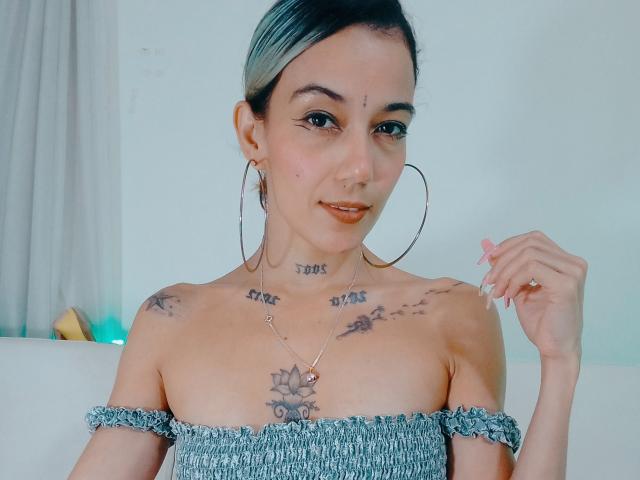 Veroo69 with dark blue hair, hoop earrings, tattoos on chest and shoulders, wearing off-shoulder gray top, smiling, hand near face, small nose ring, necklace with flower pendant.