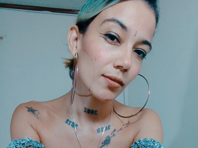 Veroo69 has blue-tipped hair, hoop earrings, tattoos on her neck and shoulders, and a cute, slightly pouty face. She's wearing an off-shoulder top with a subtle pattern.