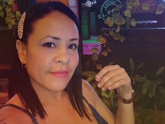 MimiP tiene la piel de un marrón medio, cabello negro y lacio con una pinza adornada con joyas, lápiz labial rosa y pequeños aretes. Lleva una blusa gris, tiene una mano levantada y está sonriendo ligeramente..