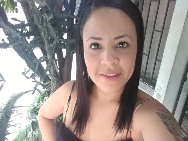 MimiP has medium brown tits, straight black hair, tan skin, and a tattoo on her right shoulder, smiling at the camera. Her green eyes are slightly upturned.