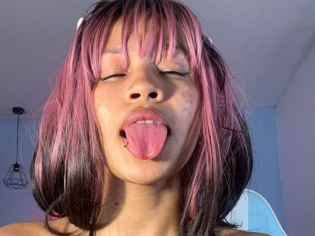 PeggyI ha i capelli rosa e neri, gli occhi chiusi, la lingua fuori, mostrando un piccolo muco sul naso, con le labbra leggermente socchiuse. Indossa una fascia bianca..