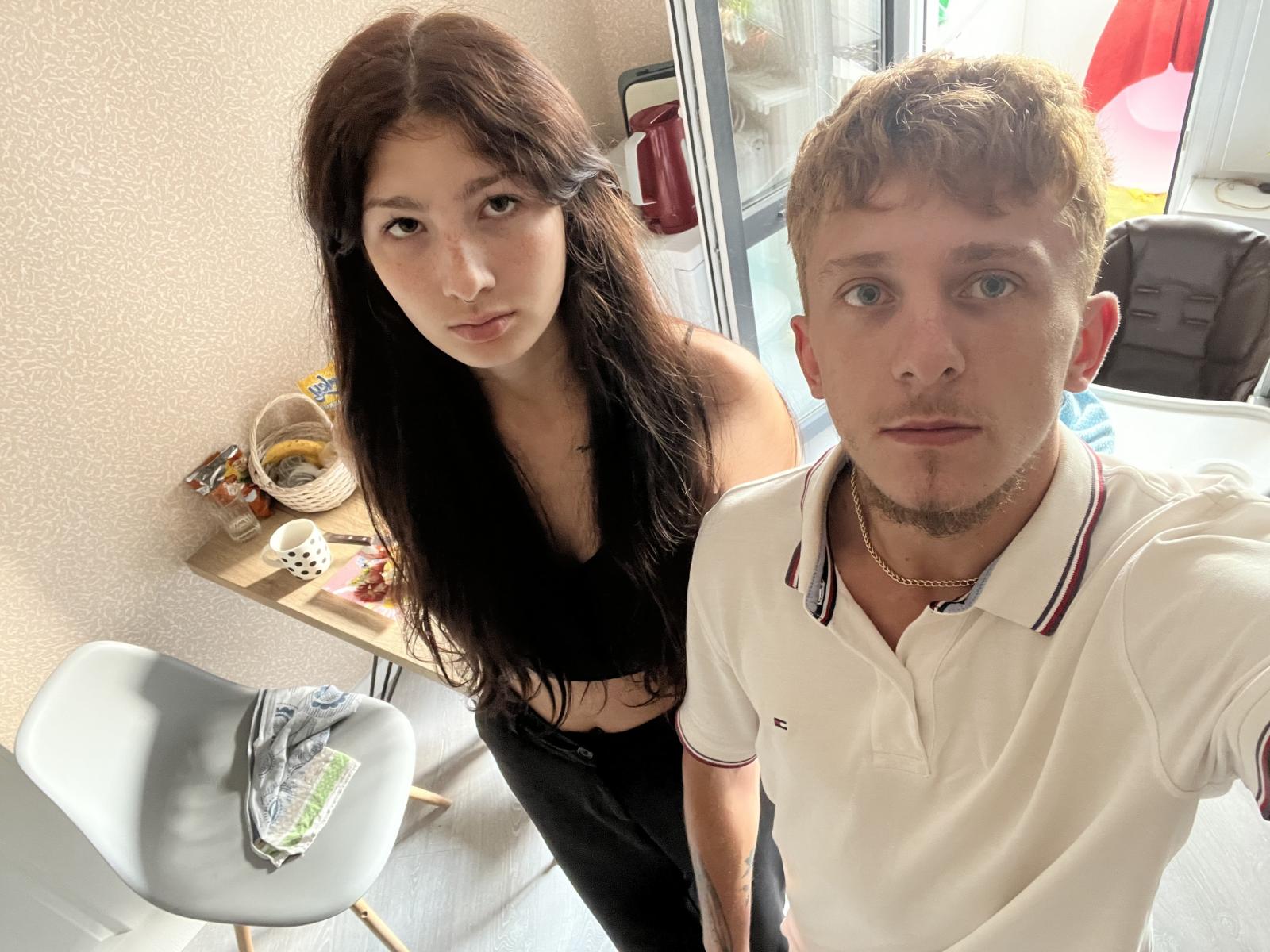 SomaAbe, met lang zwart haar en kleine borsten, gekleed in een zwarte top, staat naast SomaAbe met krullend bruin haar, een witte polo en een getatoeëerde nek, in een zonnige kamer..