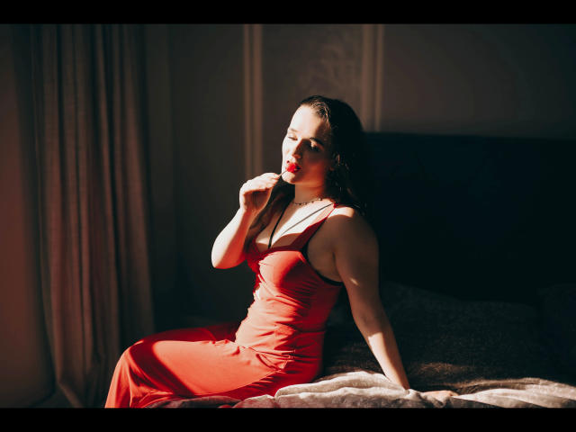 Kate-Erotic in a red dress, sitting on a bed, sunlight on her, finger in mouth, dark hair, fair skin, curvy, boobs visible, soft light, sexy, seductive.