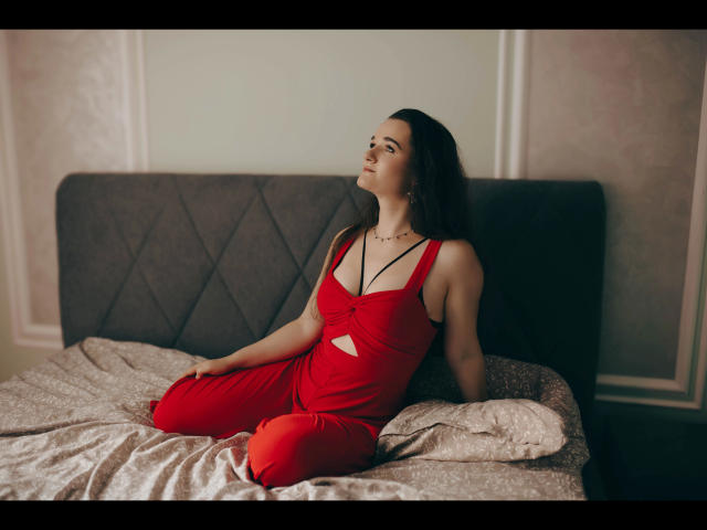 Kate-Erotic in red dress, sitting on bed, looking up, with her ass slightly raised, long dark hair, necklace, and cute face, in a soft-lit bedroom.