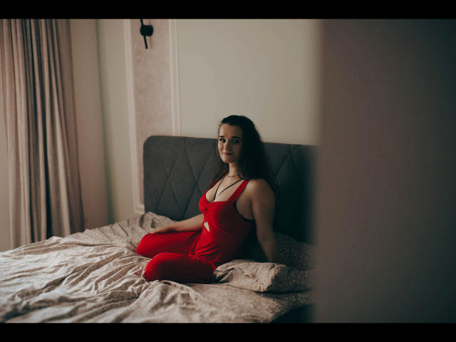 Kate-Erotic in a red dress sitting on a bed with her ass up, legs together, and looking at the camera. She has dark hair and a necklace.