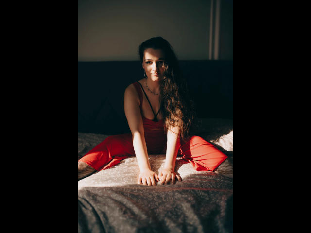 Kate-Erotic with long brown hair, wearing a red dress, crouches on a bed, hands on gray blanket, sunlight on her face, smiling, showing her ass.