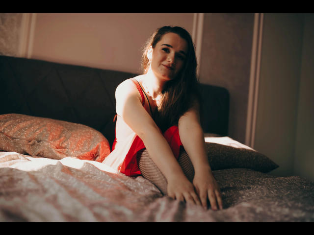 Kate-Erotic with pale skin, brown hair, and red dress, sitting on a bed with patterned sheets and pillows, wearing fishnet stockings, looking at the camera, smiling.