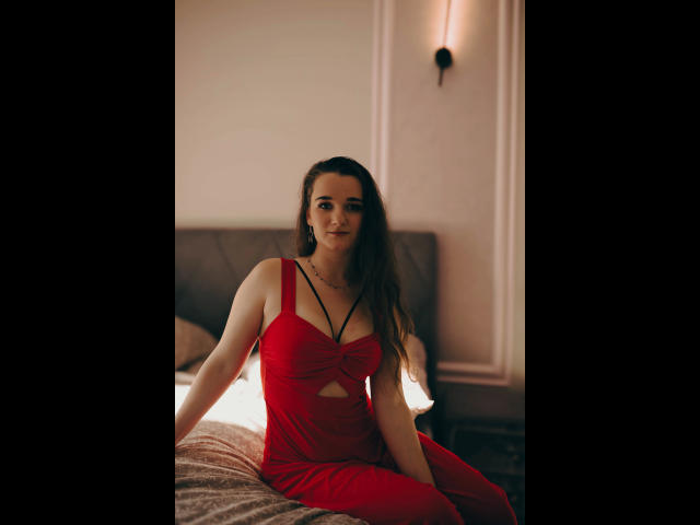 Kate-Erotic, long brown hair, wearing a red dress with a cutout, sitting on a bed, smiling, slim with small tits, fair skin, soft light, bedroom setting.