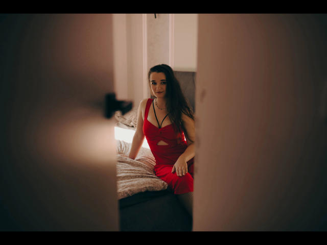 Kate-Erotic with long brown hair in a red dress sits on a bed, smiling, her tits subtly visible through the dress, seen through a partially open door.