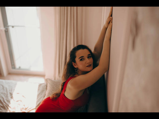Kate-Erotic with wavy brown hair, wearing a red dress, leans against a wall, arms raised, looking at camera with a slight smirk, showing her tits and soft smile.
