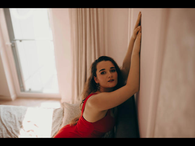 Kate-Erotic with long brown hair, red dress, and bare arms, leaning against a wall with a seductive look, her tits slightly visible, in a sunlit room.