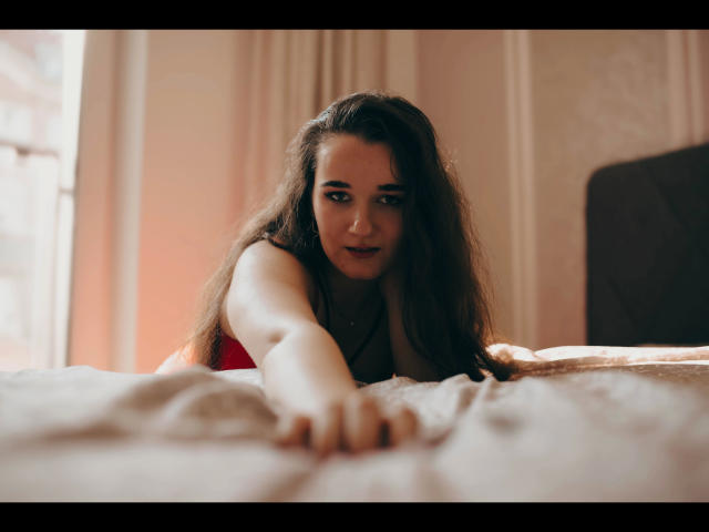 Kate-Erotic with long brown hair, looking at the camera, lying on a bed, reaching forward with her hand, wearing a red top, soft light, slightly blurred foreground.