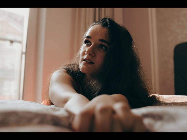 Kate-Erotic with long brown hair, lying on bed, reaching forward with hands, looking up with a slight smile, soft pink light, blurry window in background.