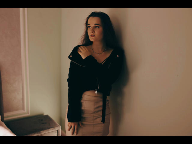 Kate-Erotic with long brown hair, black jacket, and beige skirt, standing against a wall, hands on jacket, looking to the side, subtle necklace, soft light, serious expression.