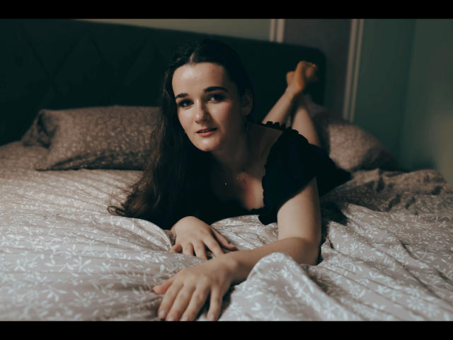 Kate-Erotic with long brown hair, black top, lying on bed, legs up, white patterned sheets, hands in front, subtle smile, bedroom setting, soft lighting, relaxed pose.