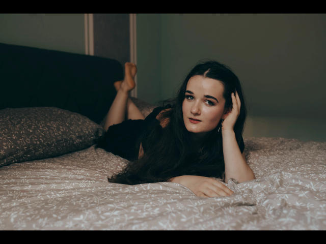 Kate-Erotic with long brown hair, lying on a bed, wearing a black shirt, legs up, arms under head, looking at camera, pale skin, subtle makeup, soft bedroom light.