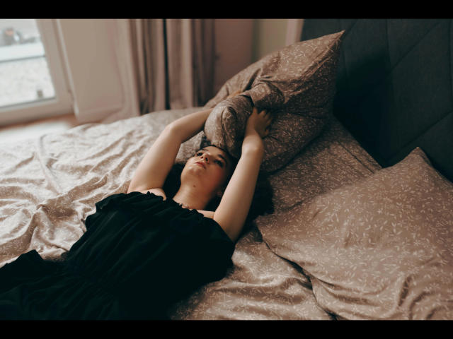Kate-Erotic lies on a bed, arms stretched above her head, holding a pillow. She's wearing a black dress, with pale skin and dark hair, in a relaxed pose.