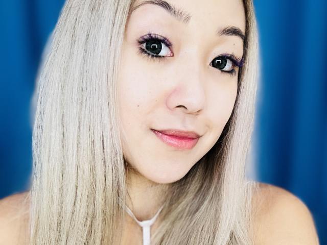 RenyLime's face with blonde hair, pink lips, and blue eyes, wearing a white necklace, looking slightly to the right with a subtle smile. Her tits are not visible.
