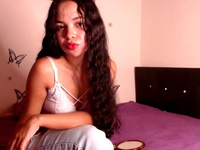 JowlinSweets, avec de longs cheveux noirs bouclés, du rouge à lèvres rouge, un débardeur en dentelle blanche et un jean bleu clair, assise sur un lit violet, regardant l'appareil photo avec un léger sourire..