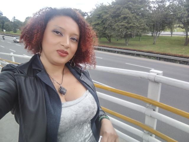 VioletaSwitch, with curly red hair, wearing a black leather jacket and white lace top, has big boobs, a necklace with a dark pendant, and a green wristband.