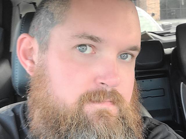 Lit with a bald head and a thick, curly brown beard, blue eyes, and a neutral expression, sitting in a car, wearing a black shirt, looking at the camera.