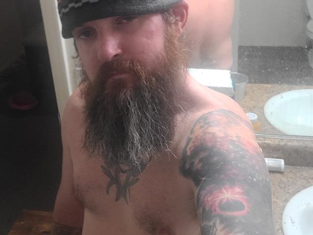 Shirtless Lit with a long, thick brown beard, wearing a gray beanie, has a large tattoo of a skull on his right arm and a black tattoo on his chest.