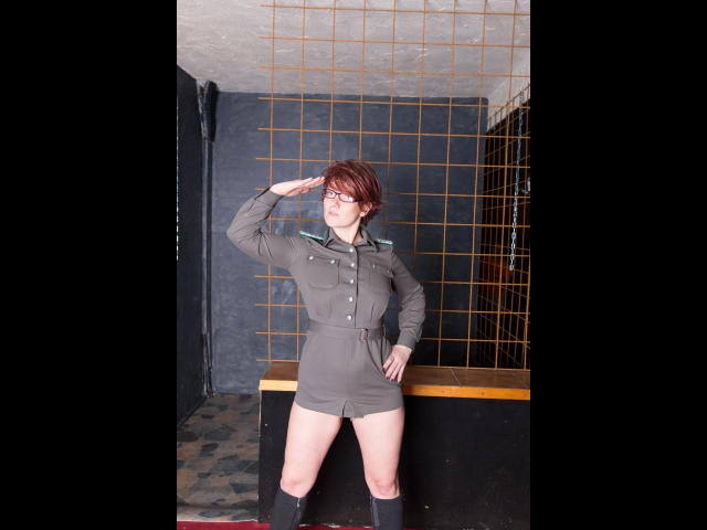 Popp-Sylvie-hot in a gray military-style dress with buttons, black knee socks, and glasses, saluting with her right hand, standing in front of a metal cage.
