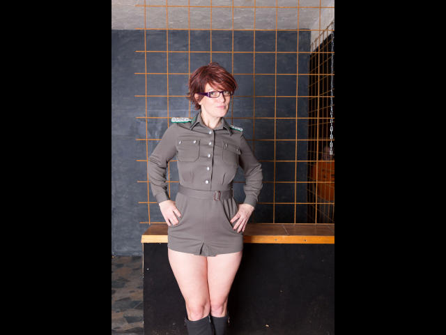 Popp-Sylvie-hot with short red hair, glasses, and a green military-style dress standing with hands on hips, showing off her ass. She's wearing black knee socks.