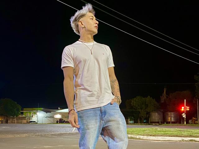 Paulo69, avec ses cheveux gris hérissés, son t-shirt blanc et son jean bleu, se tient à un carrefour nocturne, les mains dans les poches, le regard levé, avec un tatouage sur l'avant-bras gauche et un collier..