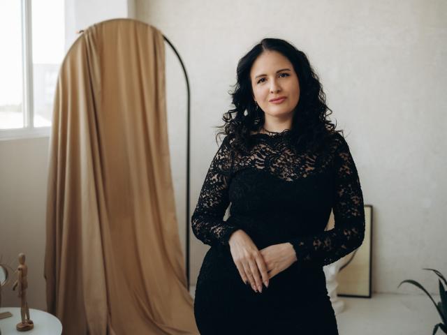 TayaBoret with curly black hair, wearing a black lace dress, standing with hands together, smiling subtly, beige curtain in the background, soft natural light.