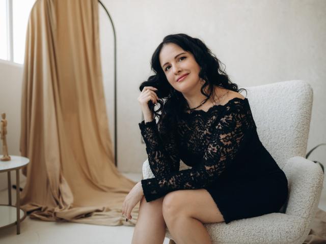 TayaBoret, com cabelos pretos encaracolados, vestindo um vestido preto de renda, sentada em uma cadeira branca, sorrindo, segurando o cabelo, mostrando seus ombros nus e suas nádegas pequenas..