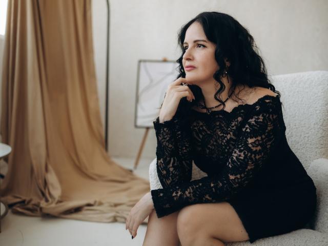 TayaBoret portant une robe noire en dentelle, assise sur une chaise blanche, la main près de la bouche, cheveux noirs bouclés, regardant sur le côté, sourire doux, yeux marrons, seins visibles sous la dentelle, rideau beige..