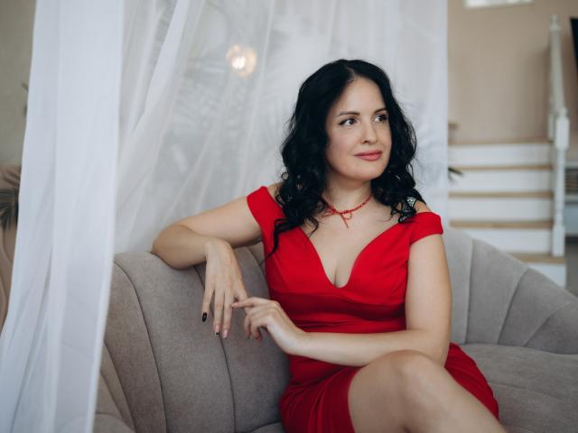 TayaBoret with black curly hair, red dress, and red necklace, sitting on a gray couch, looking to the side, right arm on the backrest, left hand on her thigh.