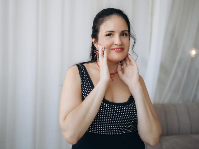TayaBoret with black hair, red earrings, and a black dress with white studs, smiling, hands near her face, showing her tits, in a soft lit indoor setting.