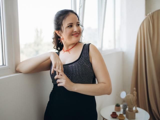 TayaBoret with curly dark hair, smiling, wearing a black polka-dot sleeveless top and black skirt, standing by a window, touching her chest, red earrings, orange necklace.