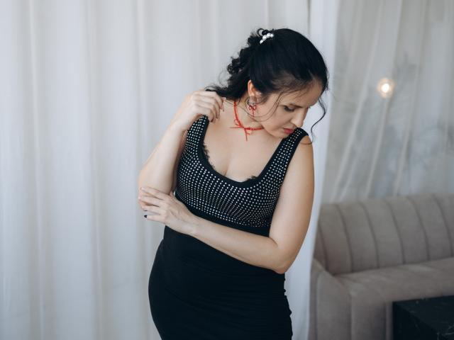 TayaBoret with dark hair in a ponytail, wearing a black polka-dot top and black skirt, touching her shoulder, red cross necklace, and red lipstick, looking down.