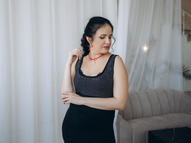 TayaBoret, curvy, with dark hair in a braid, wearing a black polka dot top and black skirt, touches red earrings, smiling, standing in a softly lit room.