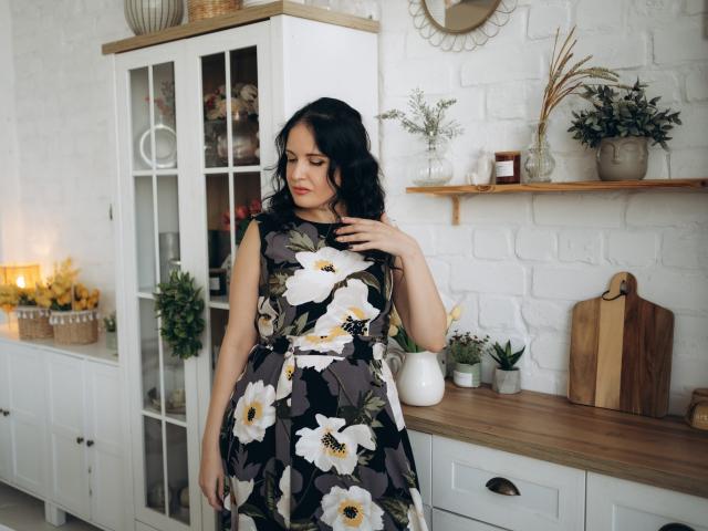 TayaBoret, con cabello oscuro y ondulado, vestida con un vestido negro con estampado floral, se toca el pecho, y está de pie en una habitación acogedora con paredes de ladrillo blanco, estanterías y armarios de madera..