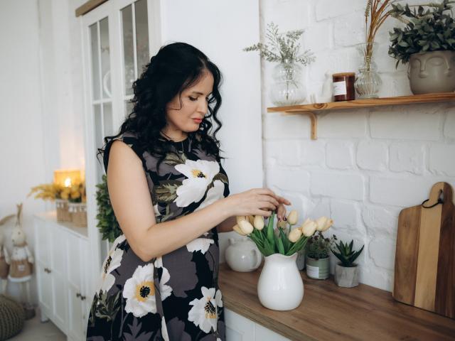 TayaBoret, con cabello negro rizado, vestida con un vestido floral, está de pie junto a una encimera de madera, sosteniendo suavemente un jarrón blanco con tulipanes amarillos. Su trasero es ligeramente visible..