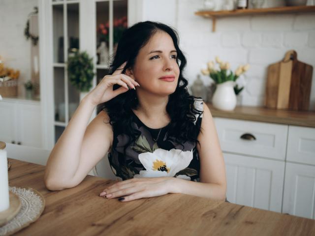 TayaBoret with black curly hair, wearing a floral sleeveless dress, sits at a wooden table, hand on chin, smiling, in a bright kitchen. Boobs are not visible.
