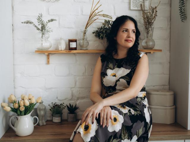 TayaBoret with black curly hair, wearing a floral dress, sitting on a wooden shelf, looking to the side, smiling, hands on knees, white brick wall, vase of tulips.