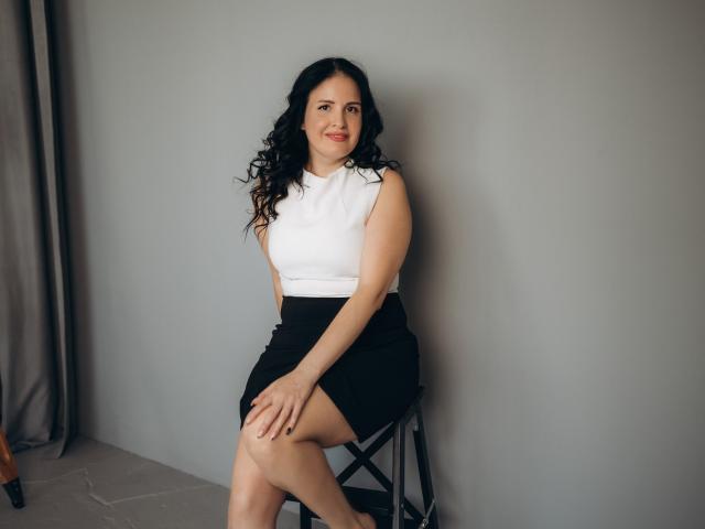 TayaBoret with curly black hair, white sleeveless top, black skirt, sitting on stool, smiling, hands on knee, gray wall, light gray curtain on left, calm pose.