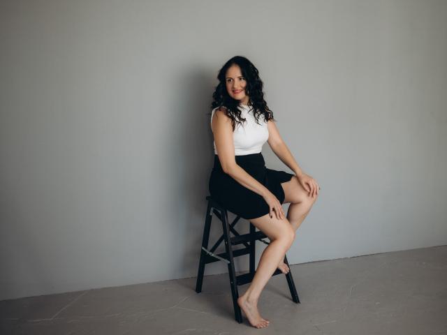 TayaBoret, with curly black hair, sits barefoot on a black stool, wearing a white sleeveless top and black skirt, smiling at the camera. Her tits are small, her ass is cute.