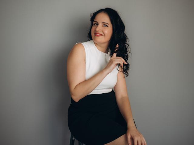 TayaBoret, com cabelo preto encaracolado, blusa branca sem mangas, saia preta, segurando o cabelo, sorrindo, sentada em um banquinho, pele clara, seios médios, fofa, confiante, fundo cinza..