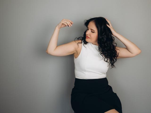 TayaBoret with curly black hair, wearing a white sleeveless top and black skirt, arms raised, touching her head, gray background, looking down, slightly smiling, pale skin.
