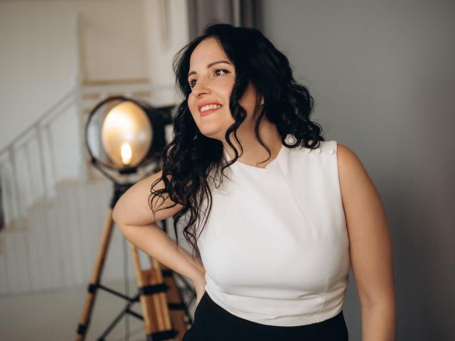 TayaBoret with curly black hair, white sleeveless top, and black skirt, smiling at camera, hand on hip, standing next to a large studio light. Her tits are average.