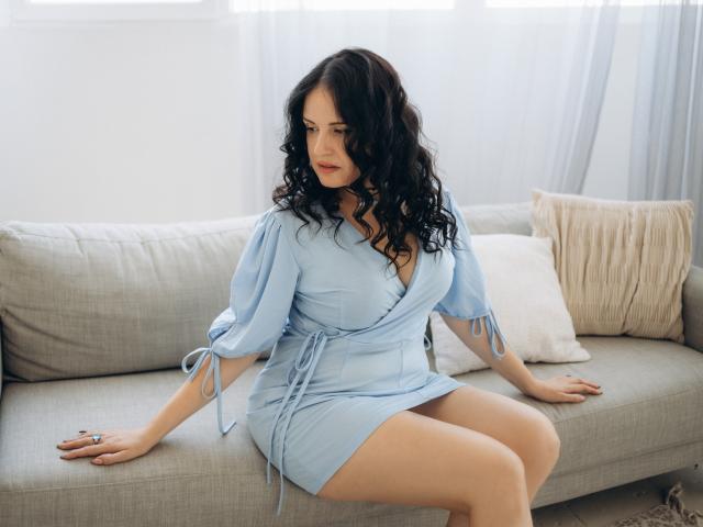 TayaBoret has curly black hair, wearing a light blue dress with tied sleeves, sitting on a gray couch, showing her ass, with a serious expression, looking down.