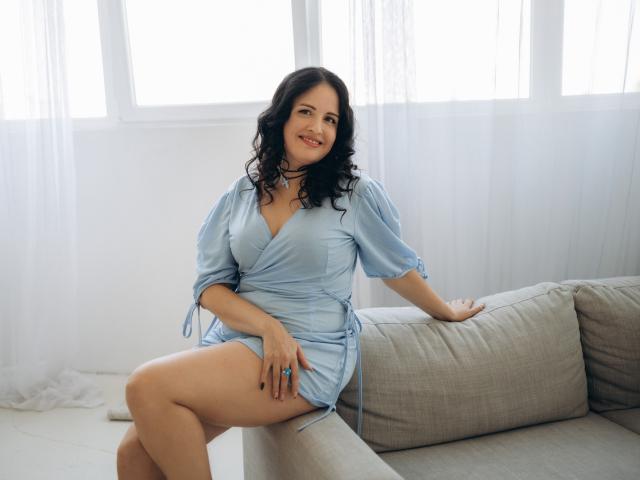 TayaBoret with curly black hair, light blue dress, sitting on a beige couch, smiling, showing her bare thighs, blue ring, white curtains, bright window, natural light.
