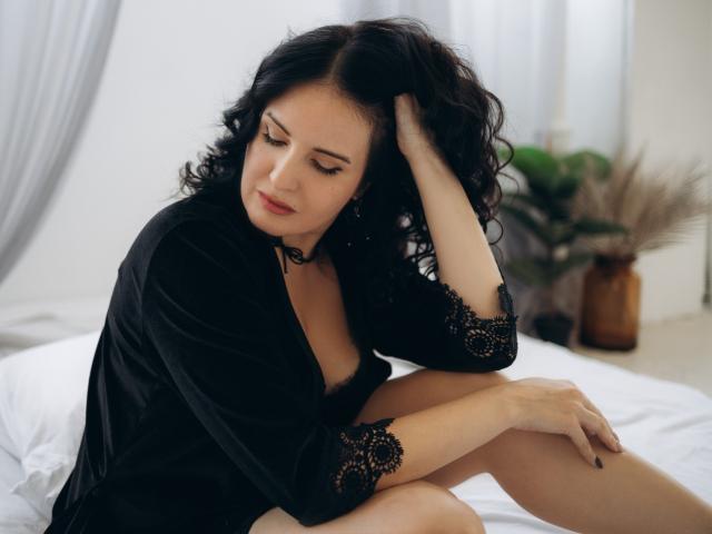 TayaBoret with curly black hair, wearing a black lace robe, sits on a bed, hand in hair, looking down, showing her bare boobs, with a slight pout.
