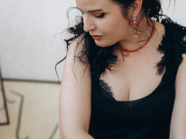 TayaBoret heeft golvend zwart haar, rode oorbellen en een zwart kanten topje dat haar borsten laat zien. Ze kijkt naar beneden met een lichte glimlach en draagt rode lippenstift..